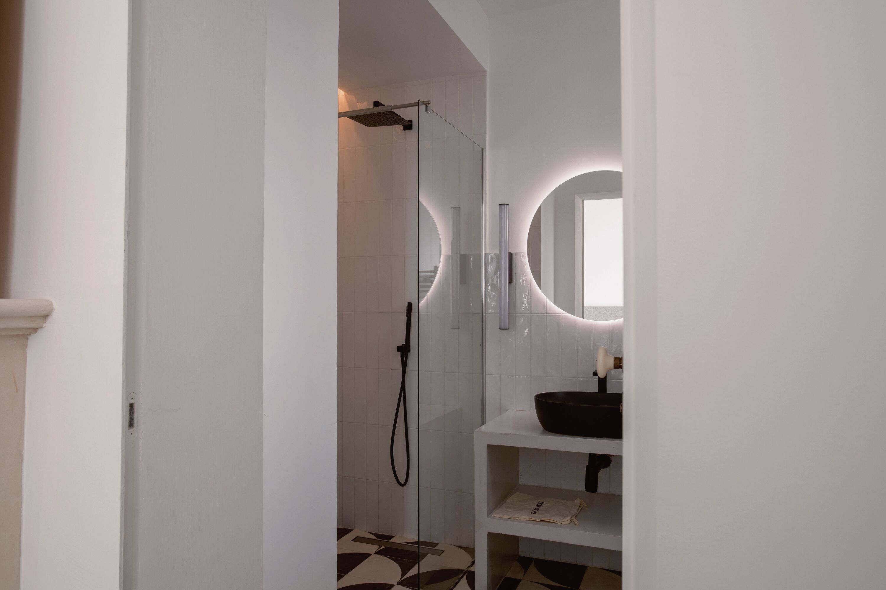 A modern bathroom with a glass-enclosed shower, a white sink with a black basin, and a mirror with a circular light.