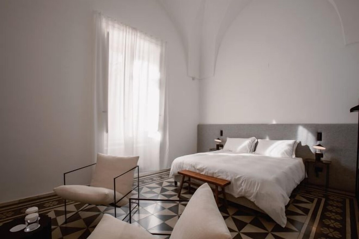A modern hotel room with a large bed, a chair, a small table, and a patterned floor.