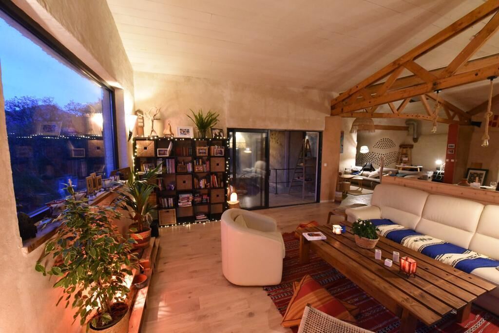 A cozy living room with a sofa, a coffee table, and a bookshelf. There is a large window on the left and a door leading to another room.