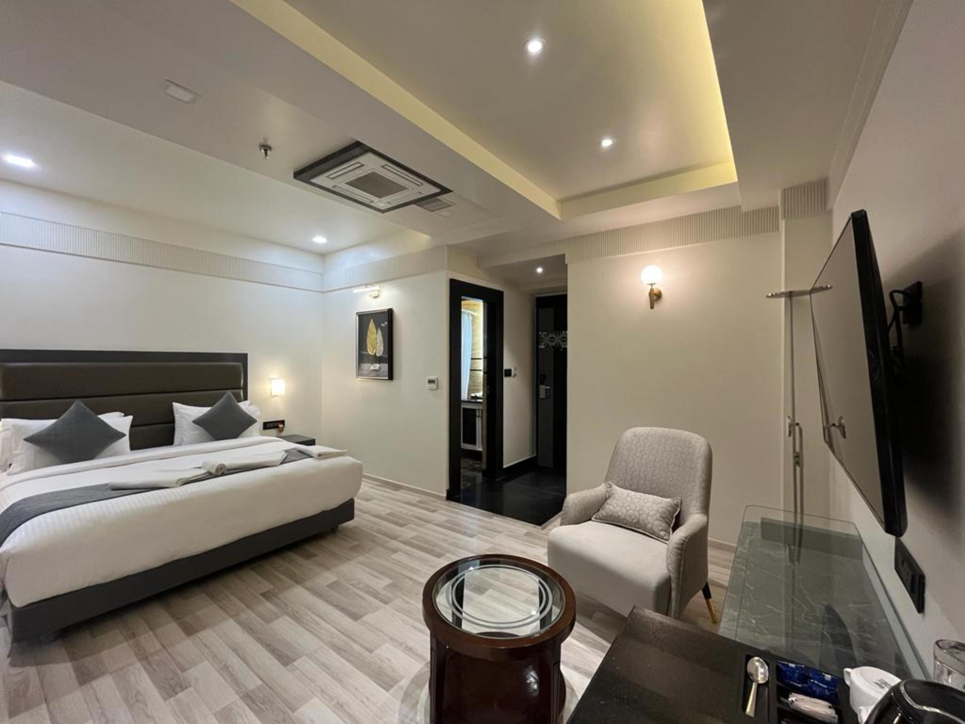 A modern hotel room with a large bed, a seating area, a glass table, and a flat-screen TV mounted on the wall.