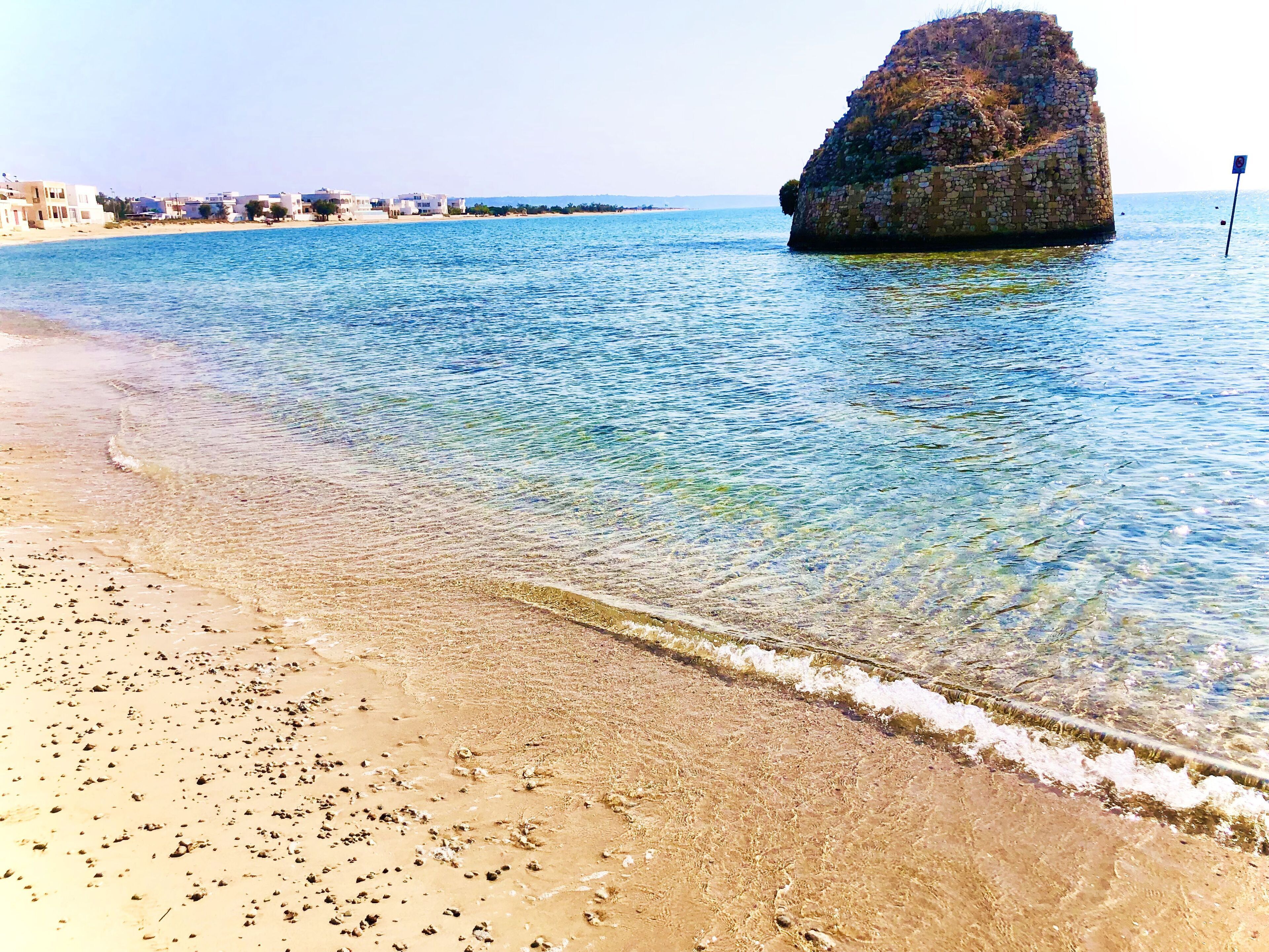 A beach with clear water, a rocky outcrop, and buildings in the background.