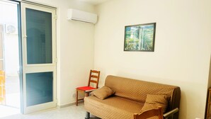 Una habitación con puerta de vidrio corrediza, un sofá color beige acolchado, una silla de madera y un cuadro en la pared.