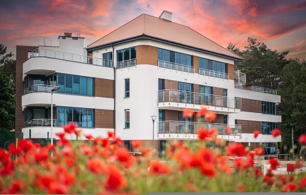 Un edificio de varios pisos con balcones y ventanas amplias, rodeado de vegetación y flores rojas.