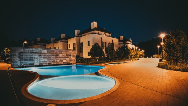 A nighttime view of a swimming pool area with a curved pool, a brick wall, and a building with multiple chimneys in the background. - Jardin Taras z Ogrodem Nowoczesny Blisko Morza (Trzebiatow)