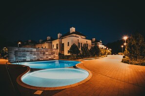 A nighttime view of a swimming pool area with a curved pool, a brick wall, and a building with multiple chimneys in the background. - Jardin Taras z Ogrodem Nowoczesny Blisko Morza (Trzebiatow)