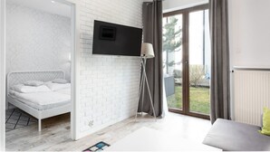 A modern bedroom with a bed, a TV mounted on a brick wall, a floor lamp, and a view of the outdoors through a glass door. - Jardin Taras z Ogrodem Nowoczesny Blisko Morza (Trzebiatow)