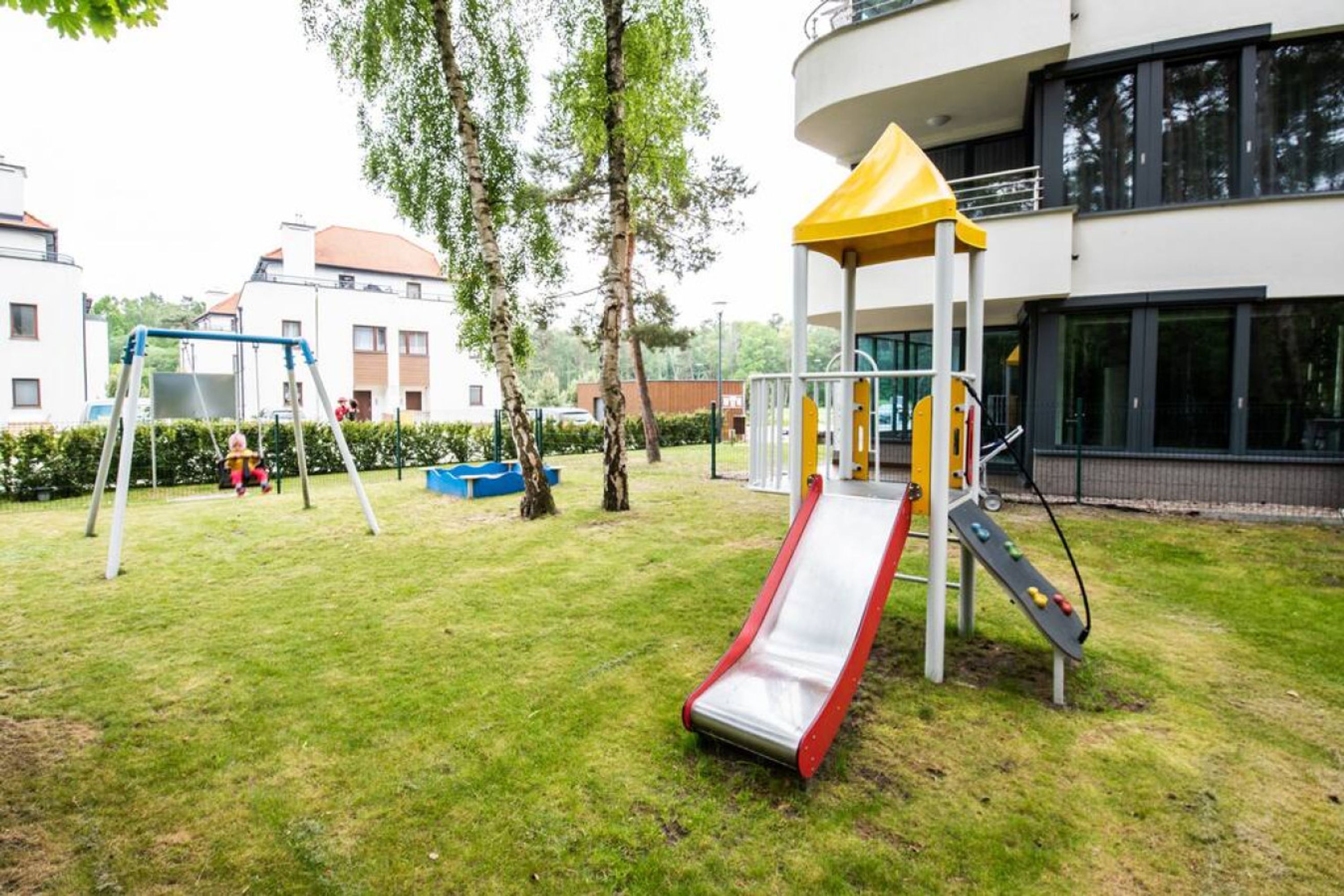 A playground with a slide and swings, surrounded by grass and trees, with buildings in the background.