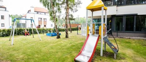 A playground with a slide and swings, surrounded by grass and trees, with buildings in the background.