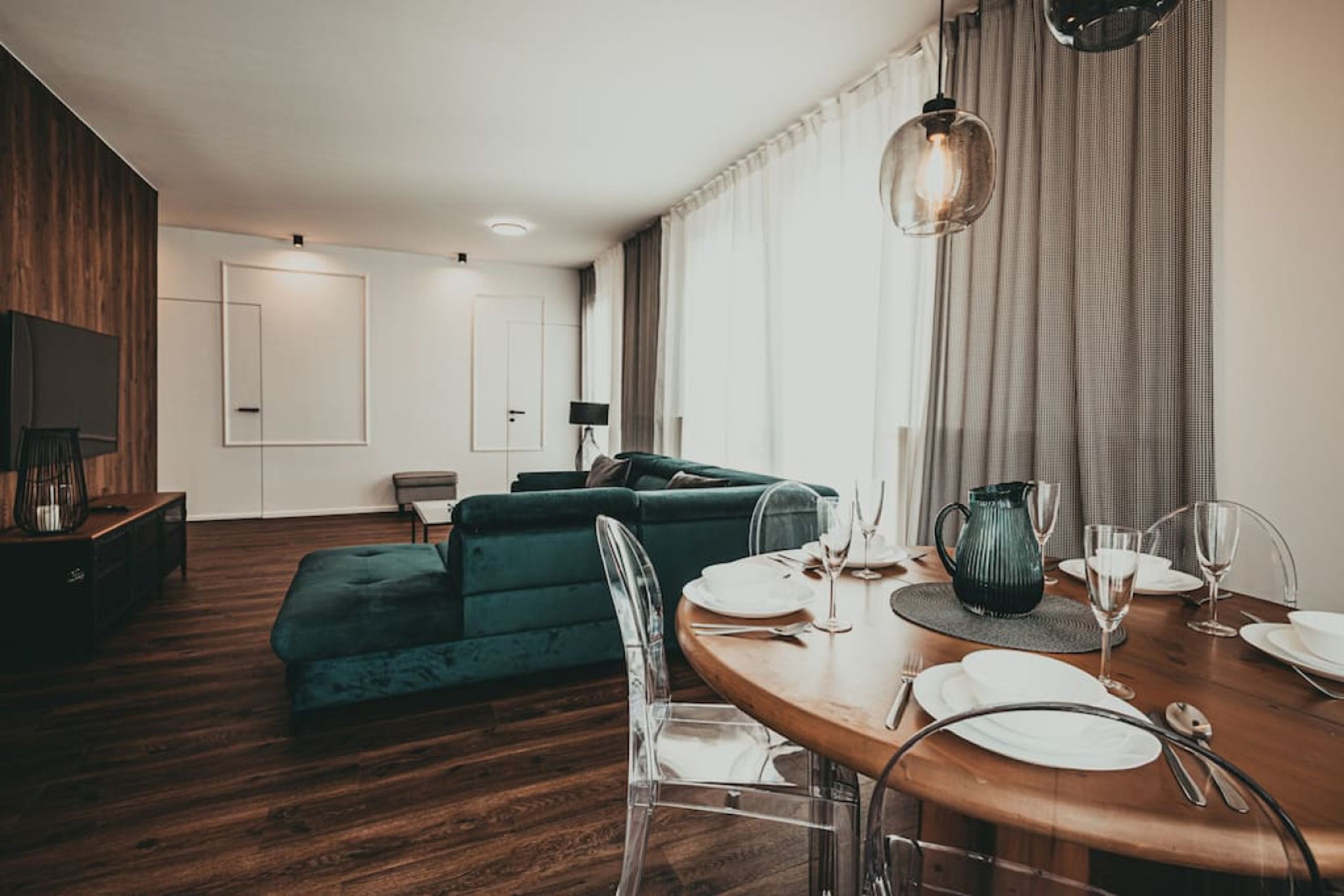 A modern living room with a wooden floor, a green sofa, a dining table set with white plates and glasses, and a TV mounted on the wall.