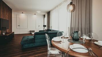 A modern living room with a wooden floor, a green sofa, a dining table set with white plates and glasses, and a TV mounted on the wall.