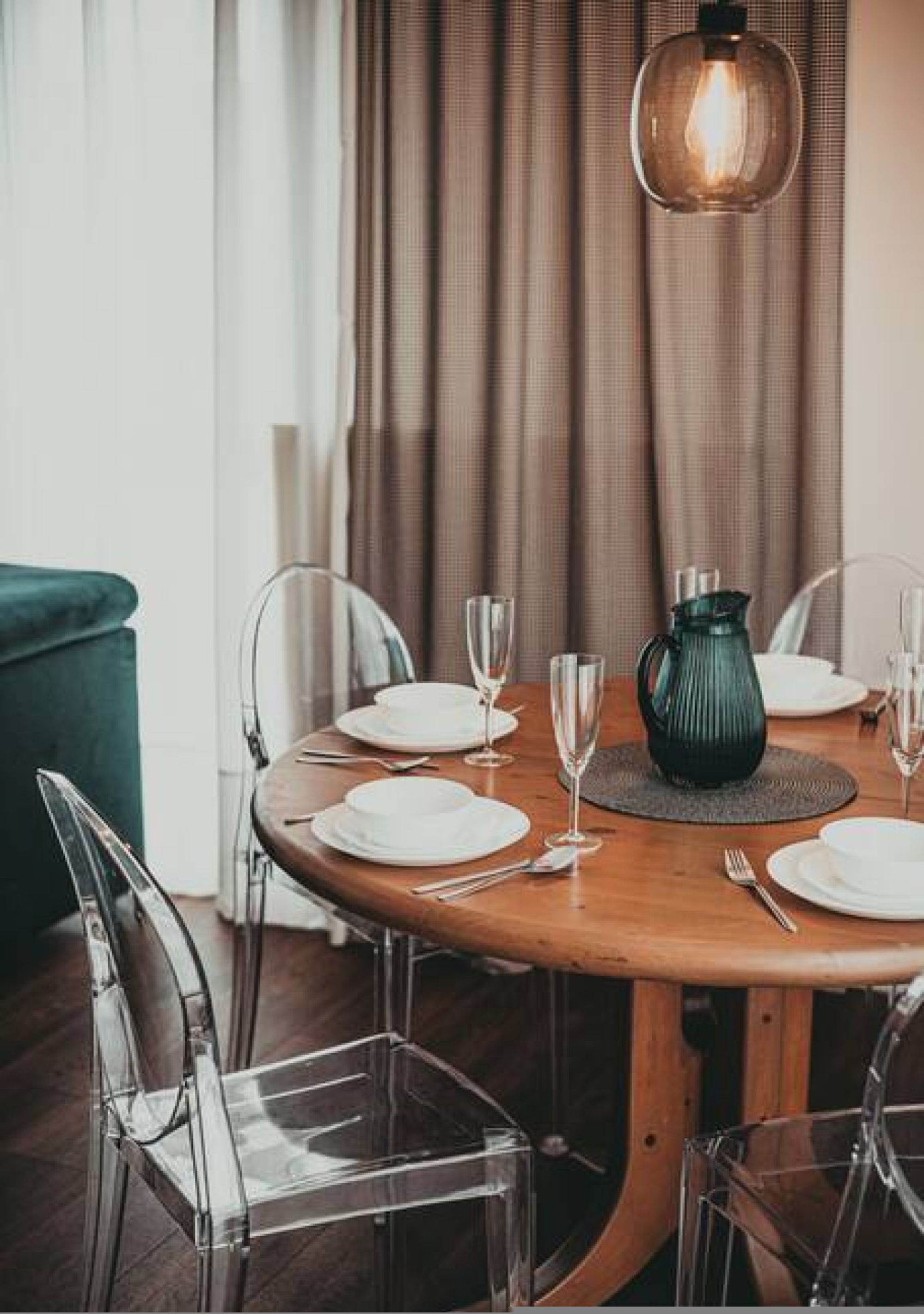 Un coin repas avec une table ronde en bois, dressée pour quatre personnes, entourée de chaises transparentes. On peut voir un pichet vert et un luminaire suspendu.