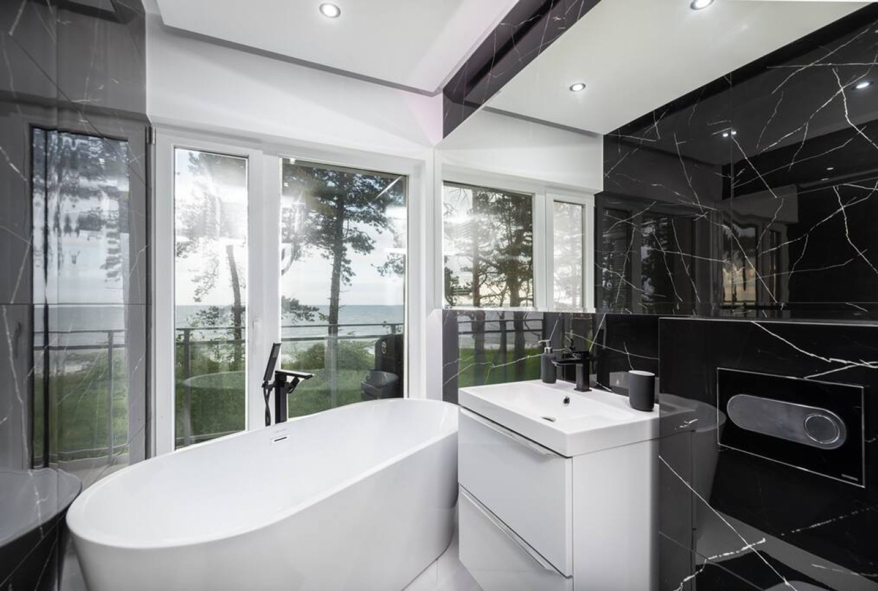 Un bagno moderno con una vasca da bagno indipendente, un mobile vanitario bianco e ampie finestre che si affacciano su alberi e uno specchio d'acqua.