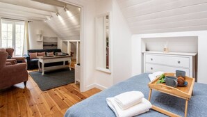 Une pièce spacieuse avec un grand lit, un coin salon comprenant un canapé et des fauteuils, une table basse en bois, et une petite kitchenette à l’arrière-plan.