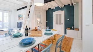 Ein Essbereich mit einem weißen Tisch, gedeckt mit blauen Tellern, Weingläsern und einer Flasche. Ein Holzkasten mit Flaschen und eine Leiter, die zu einem Schlafzimmer darüber führen.