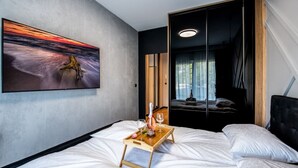 A modern hotel room with a large bed, a bedside table with a tray, a painting of a sunset, and a view into a dark room with a bed. - Hermoso Wypoczynek w Strefie SPA Blisko Morza (Mielno)
