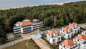 A multi-story hotel with a swimming pool, surrounded by a forested area and a beach.