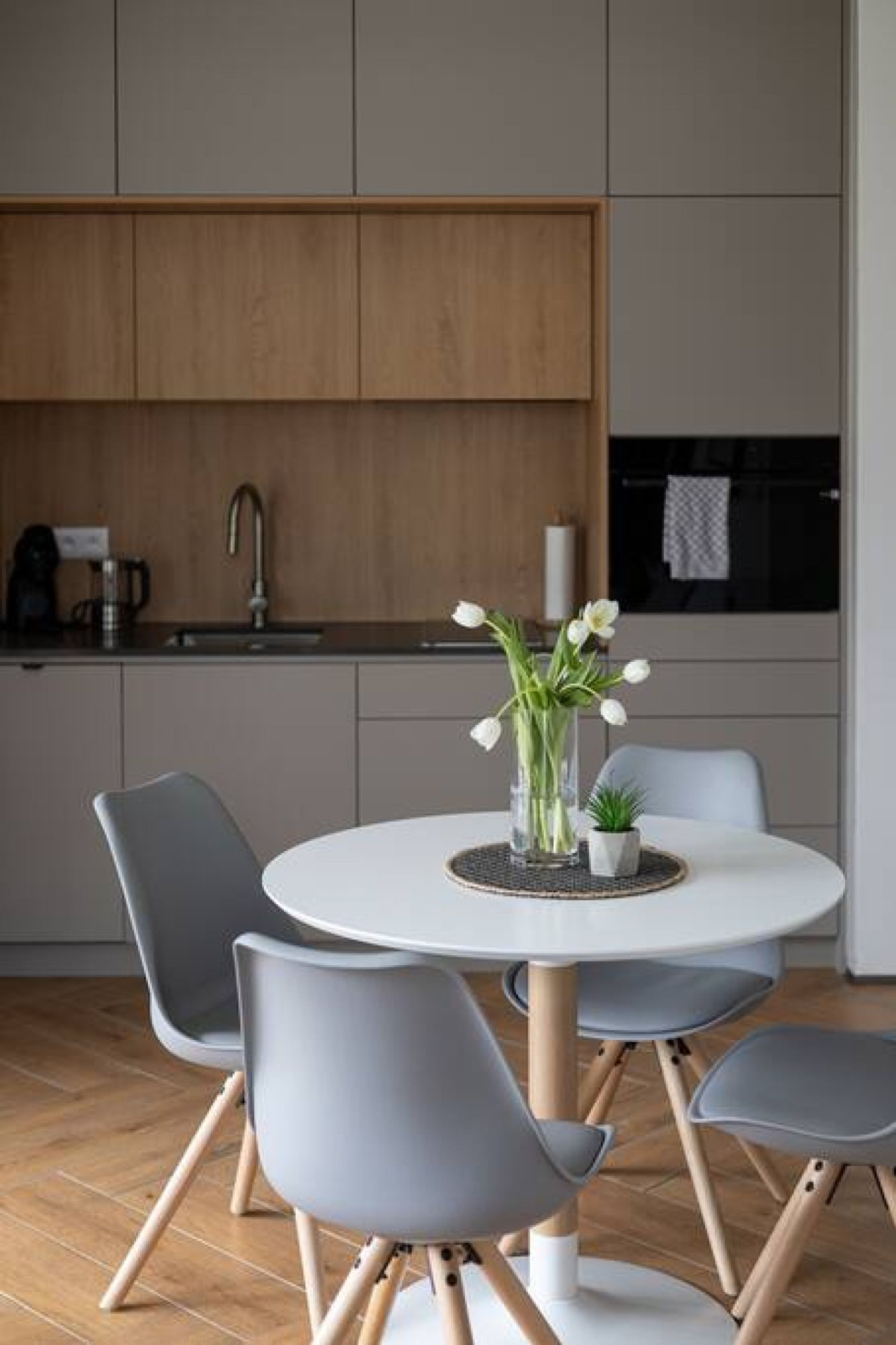 A modern kitchen with a round dining table, grey chairs, and a vase with flowers.