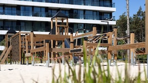 Un terrain de jeux avec des structures et des toboggans en bois, situé devant un bâtiment moderne à plusieurs étages et doté de grandes fenêtres.