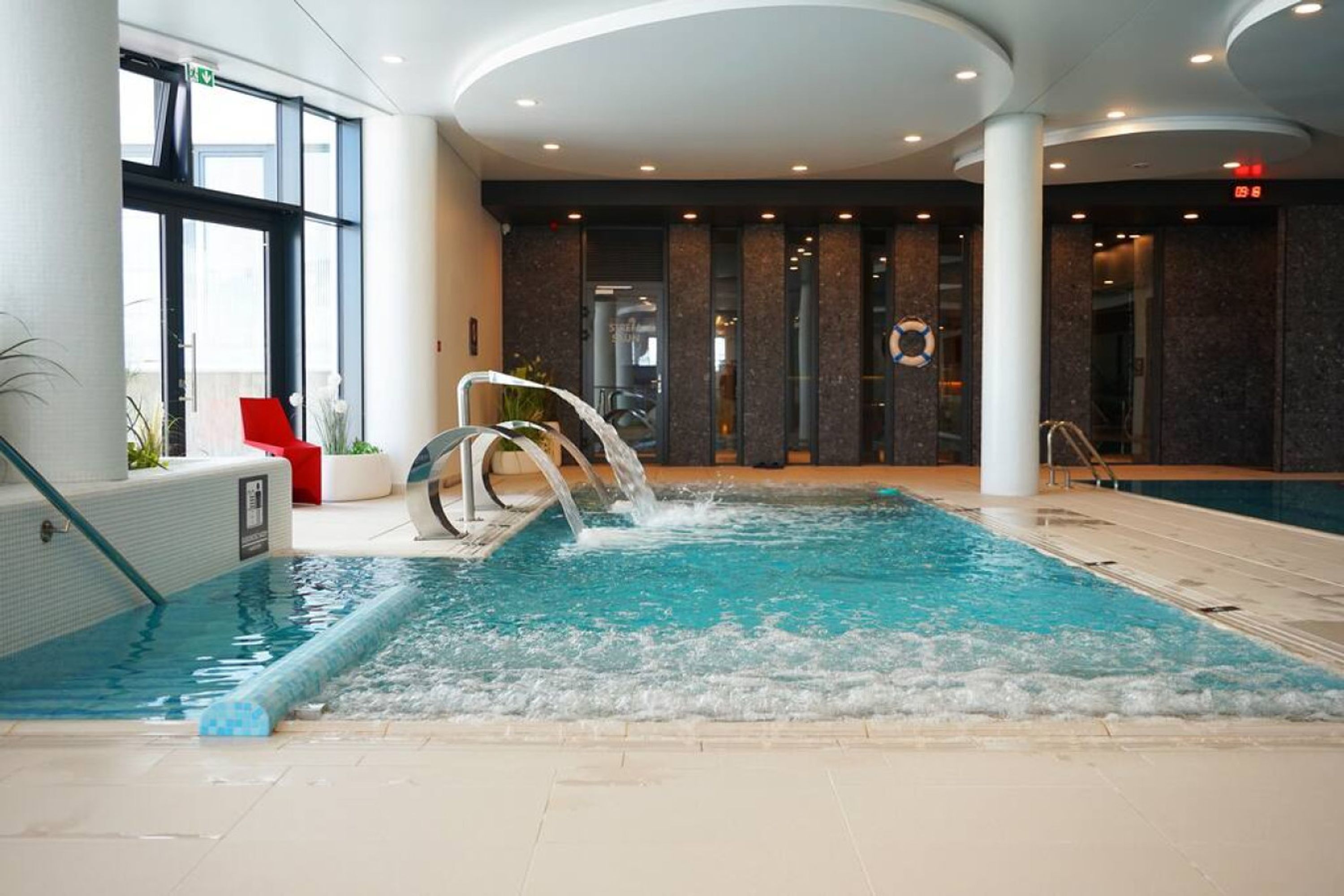 An indoor swimming pool with a water feature, surrounded by a modern interior with large windows and a seating area.