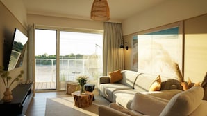 A spacious living room with a large sofa, a coffee table, and a television. There is a sliding glass door leading to a balcony with a view of a body of water and greenery. - View Luksusowy Apartament SPA Ko Obrzeg Polanki Aqua (Kołobrzeg)