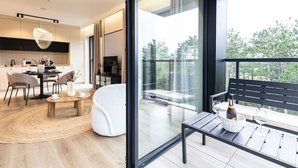 A modern living room with a glass wall, a dining area with a round table, and a balcony with a bench. - Sunrise Tw j Prywatny Widok na Ba tyk (Trzebiatow)