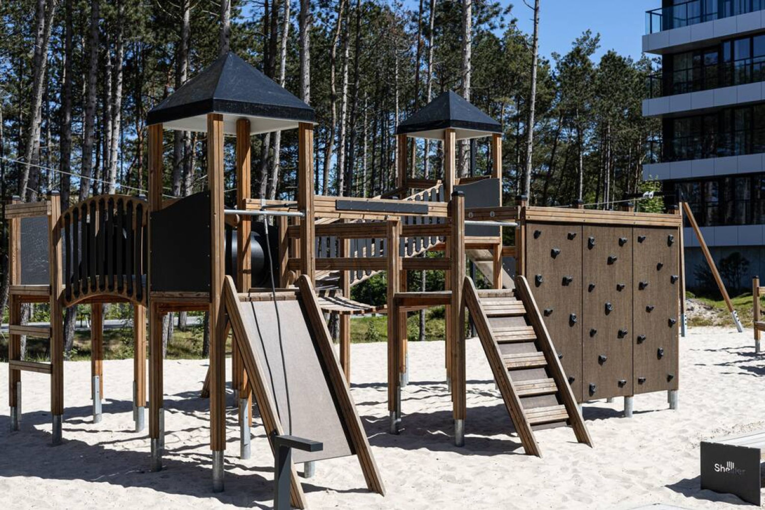 A wooden playground structure with slides and climbing areas, surrounded by sand and trees.