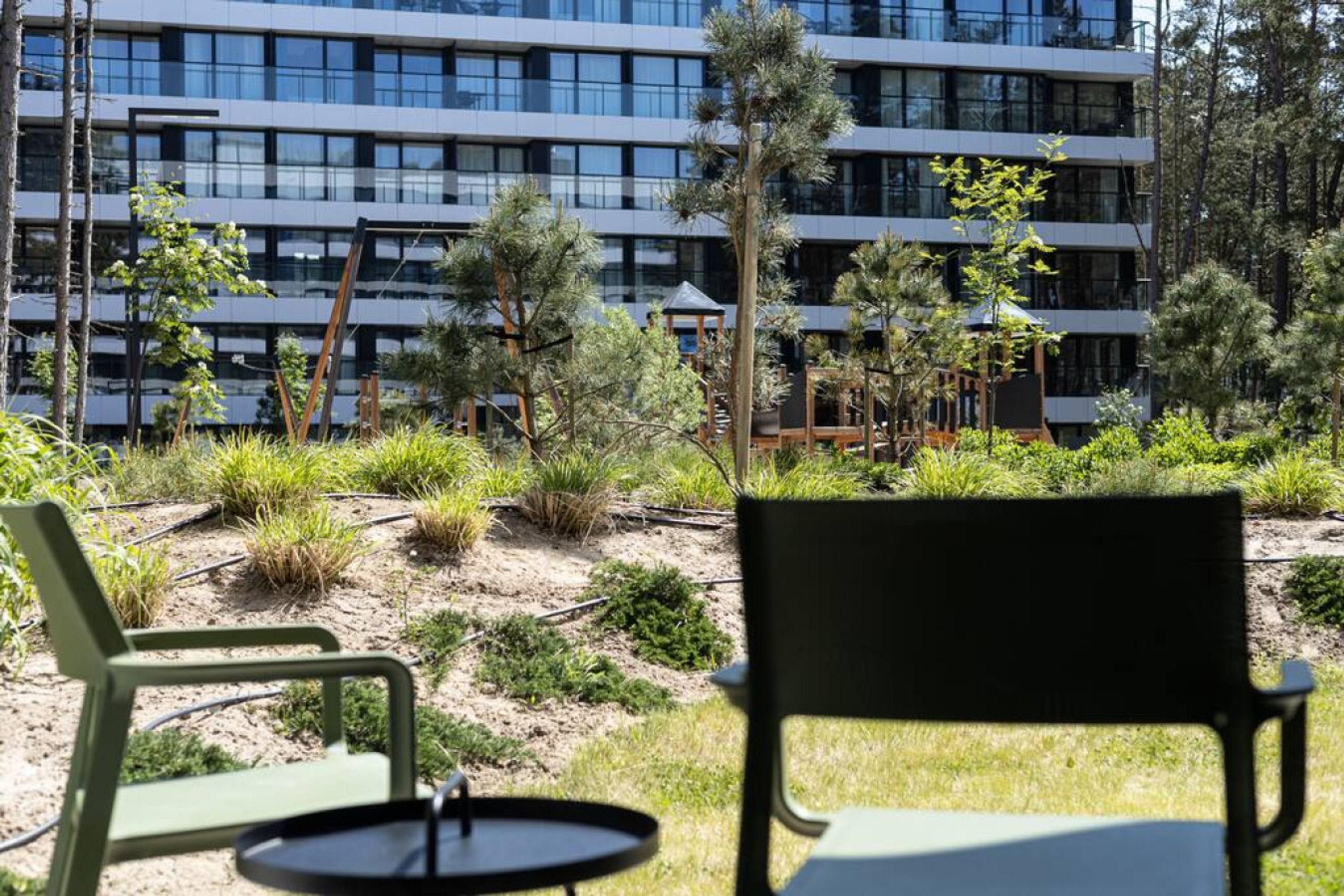 An outdoor seating area with chairs and a table, surrounded by greenery and a modern building in the background.