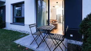 Ein modernes Haus mit Terrasse, ausgestattet mit einem Holztisch und -stühlen, einer Schiebetür zu einer Küche und einem kleinen Garten.