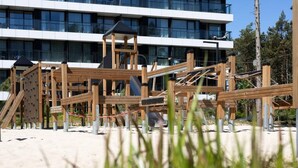 Een speeltoestel met een houten glijbaan en klimelementen, met op de achtergrond een modern gebouw met meerdere ramen.