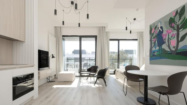 A modern living room with a flat-screen TV, a dining area with a round table and chairs, and large windows offering a city view. - Art Forrest 33 M in Rogowo (Trzebiatow)