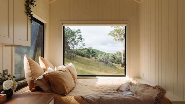 Ein Schlafzimmer mit großem Fenster, einem Bett mit beigefarbener Bettwäsche und einem Nachttisch aus Holz mit Blumen in einer Vase.