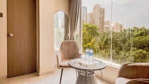 Una habitación con puerta, una mesa redonda con sillas, una ventana con cortinas y vistas a edificios y zonas verdes.