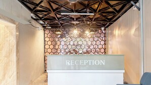 Mostrador de recepción con el rótulo 'RECEPTION', una pared con diseño hexagonal en el fondo, techo con estructura de armadura metálica y una silla azul a la derecha.