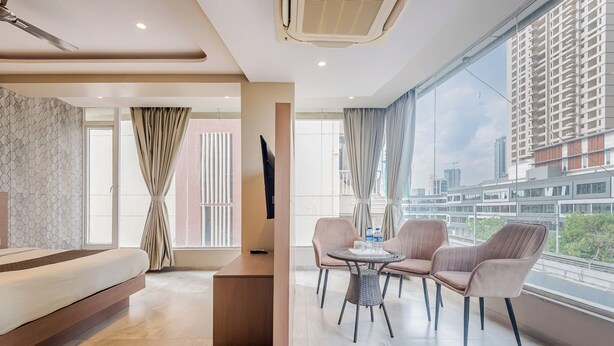 Habitación de hotel moderna con cama, una pequeña mesa con sillas, televisión y ventanas amplias con vistas a la ciudad.