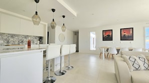 A modern kitchen with white cabinets, a marble backsplash, and pendant lighting. There is a dining area with a white table and chairs, and a sofa with a patterned throw pillow. - Belle Maison 4 ch- 8pers. Mornac (Mornac-sur-Seudre)