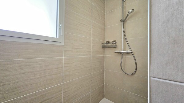 Une salle de bain avec des carreaux de couleur beige, une douche équipée d’un pommeau de douche à main et une fenêtre.