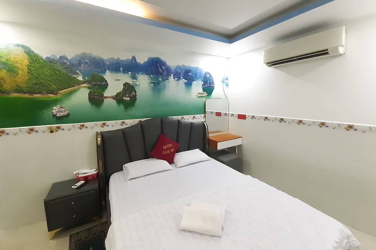 A hotel room with a bed, a nightstand, a small table, and a wall mural depicting a scenic landscape.