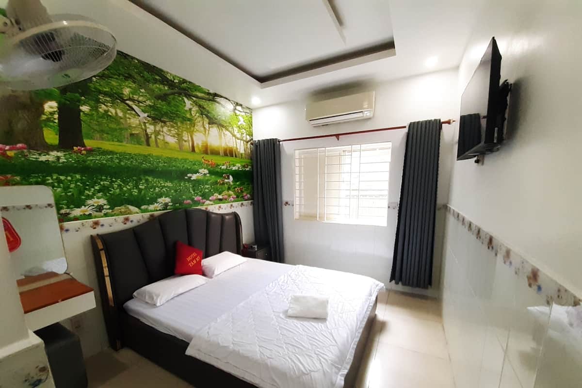 A hotel room with a bed, a wall-mounted TV, an air conditioning unit, a window with curtains, and a mural of a park scene.