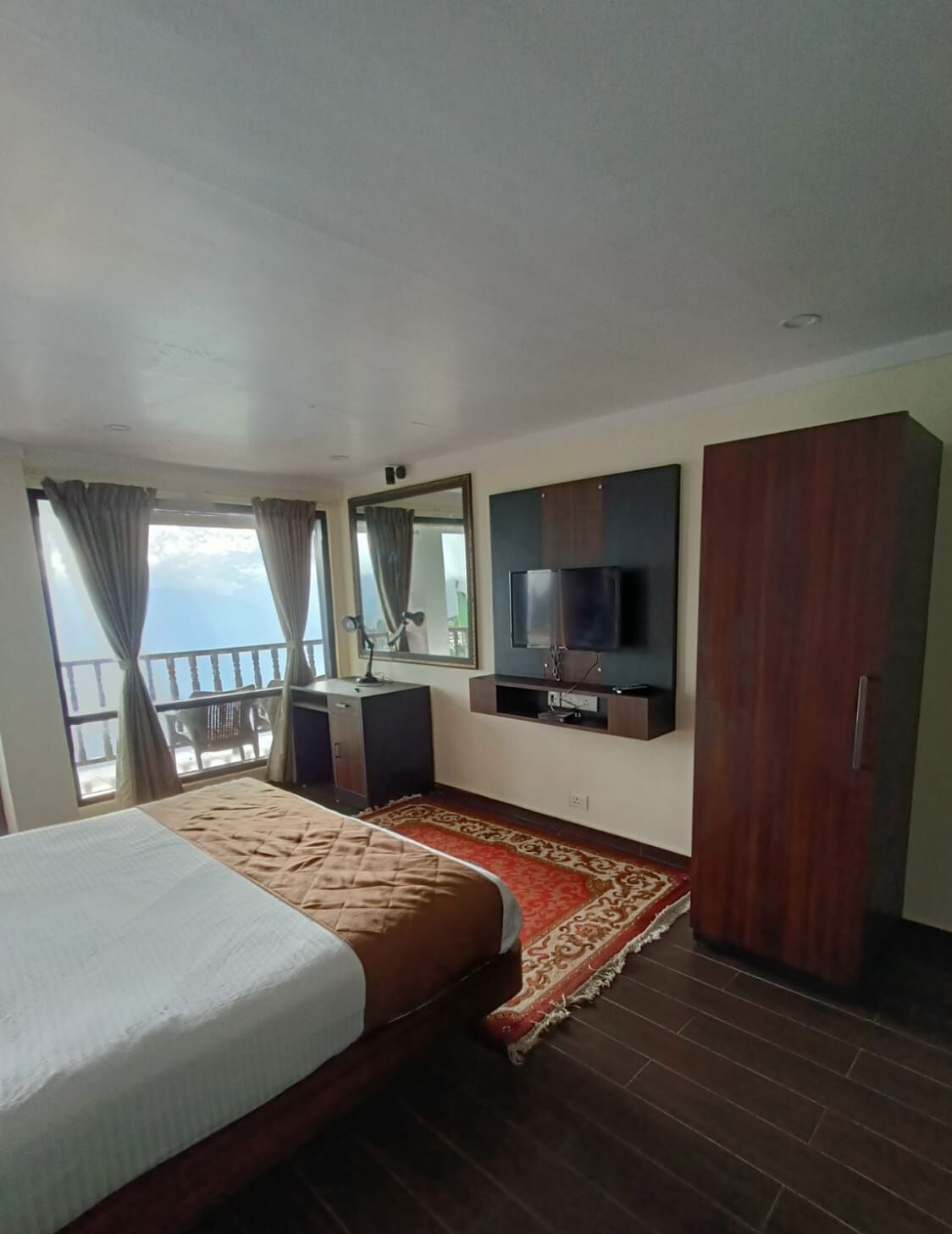A hotel room with a large bed, a TV mounted on the wall, a wardrobe, a desk with a chair, and a balcony with curtains.