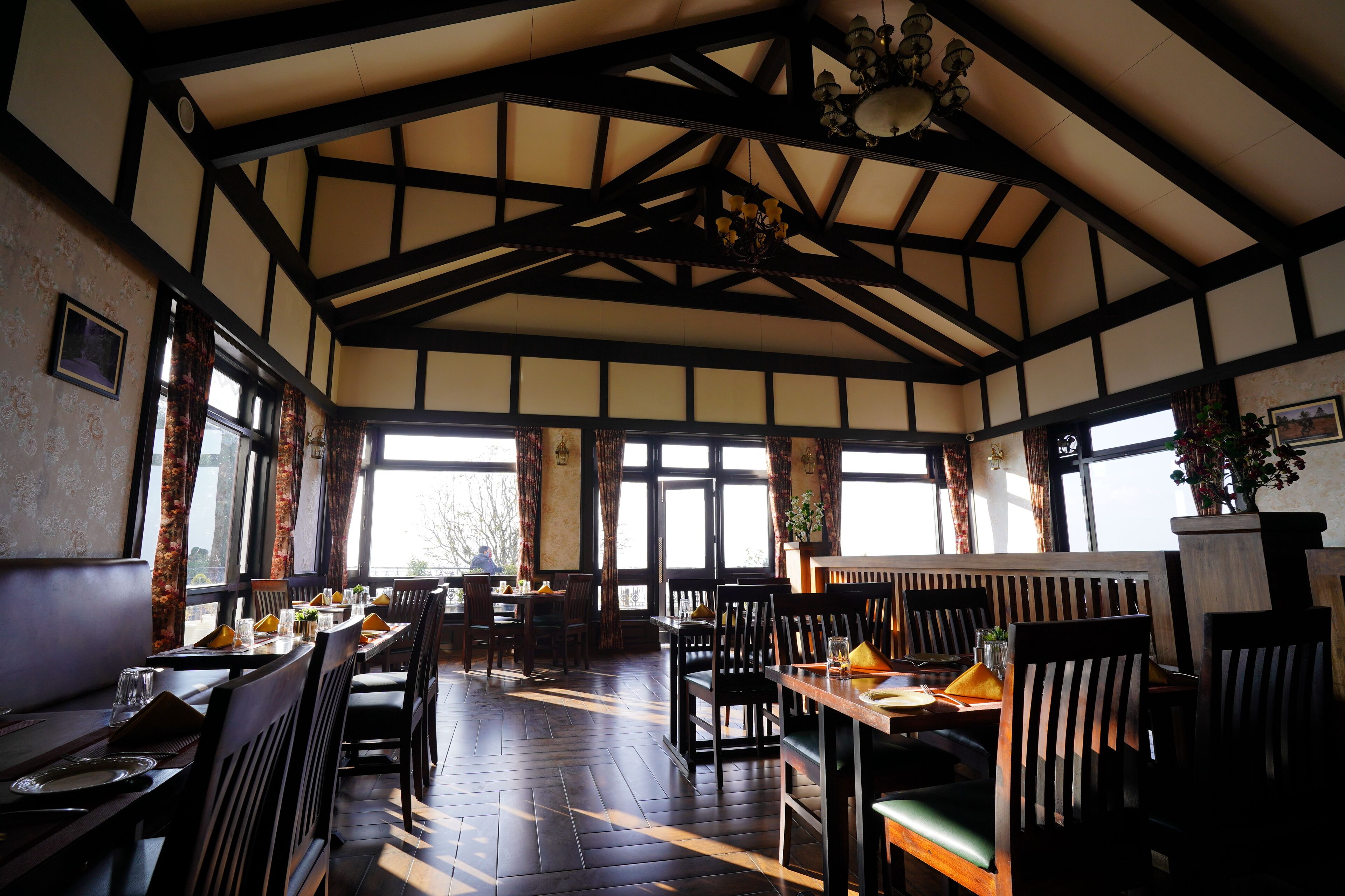 A restaurant with high ceilings, exposed beams, and large windows. Tables are set with chairs, and there are plants on the tables.