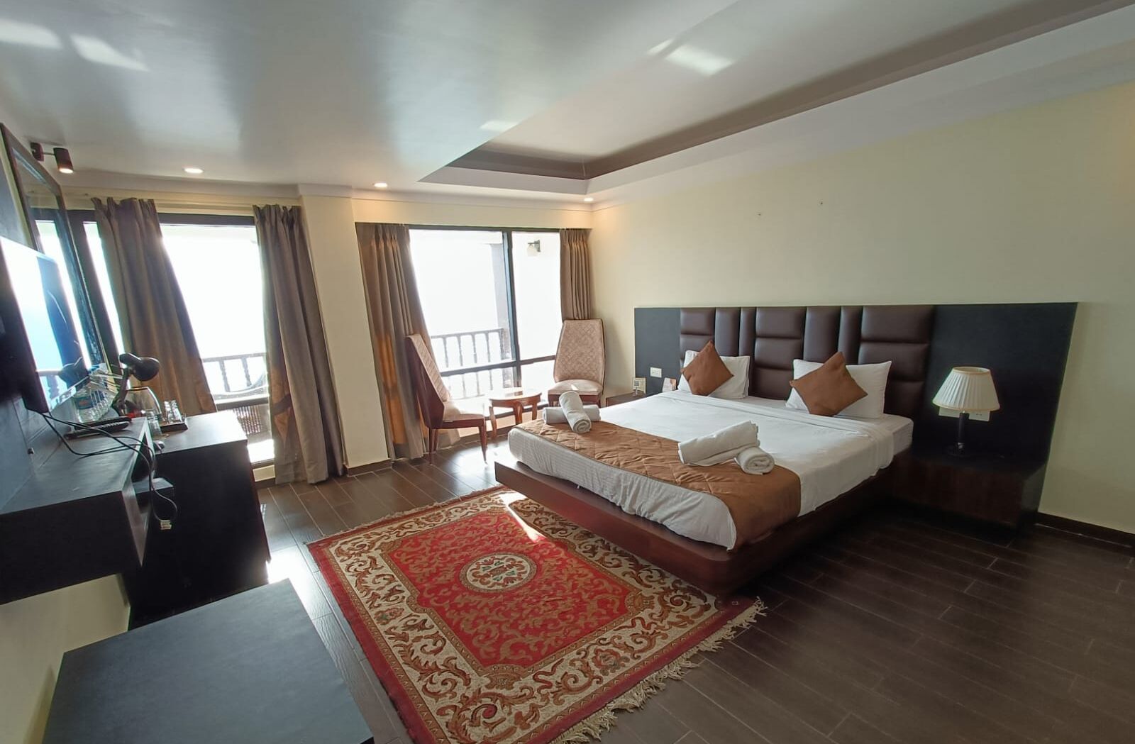 A hotel room with a large bed, a desk, a chair, a nightstand with a lamp, and a view of the outdoors.