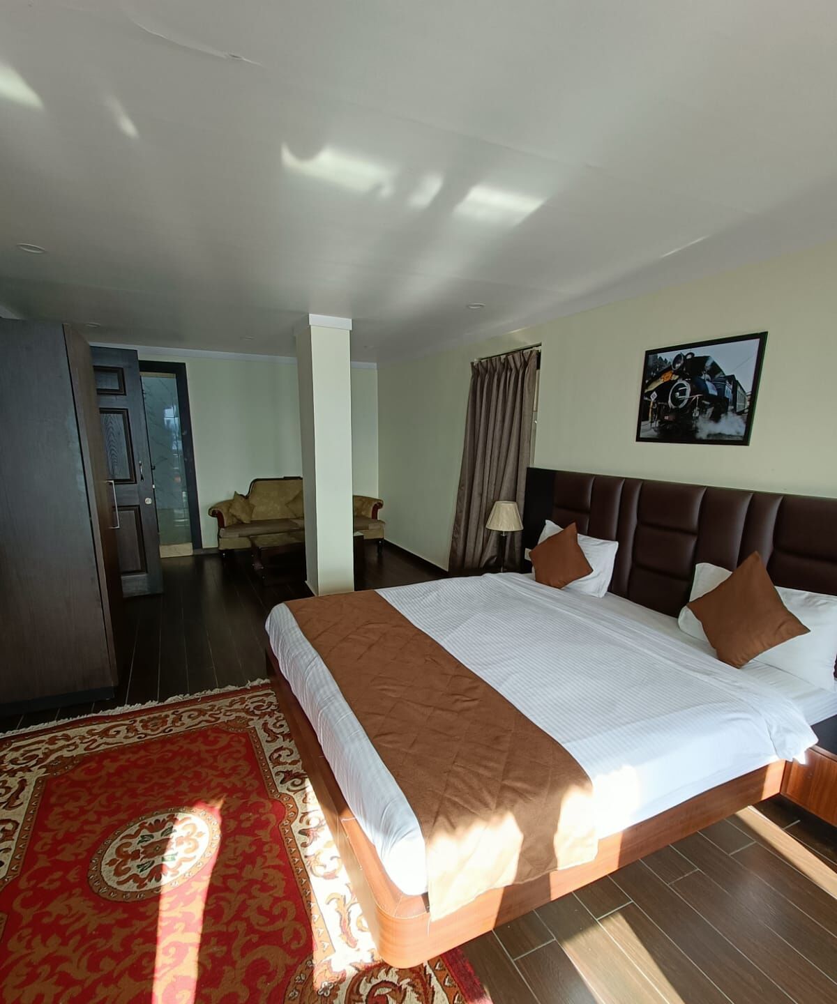 Habitación de hotel con una cama grande, un tapete rojo estampado, un armario de madera y una puerta que da a otra habitación.