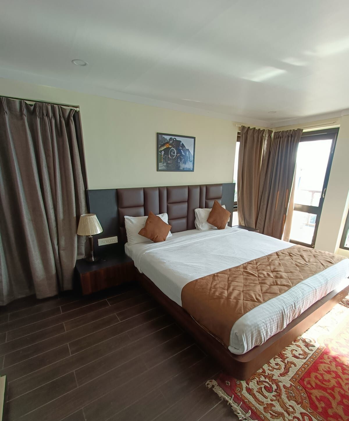 A hotel room with a large bed, wooden headboard, brown curtains, a nightstand with a lamp, and a painting on the wall.