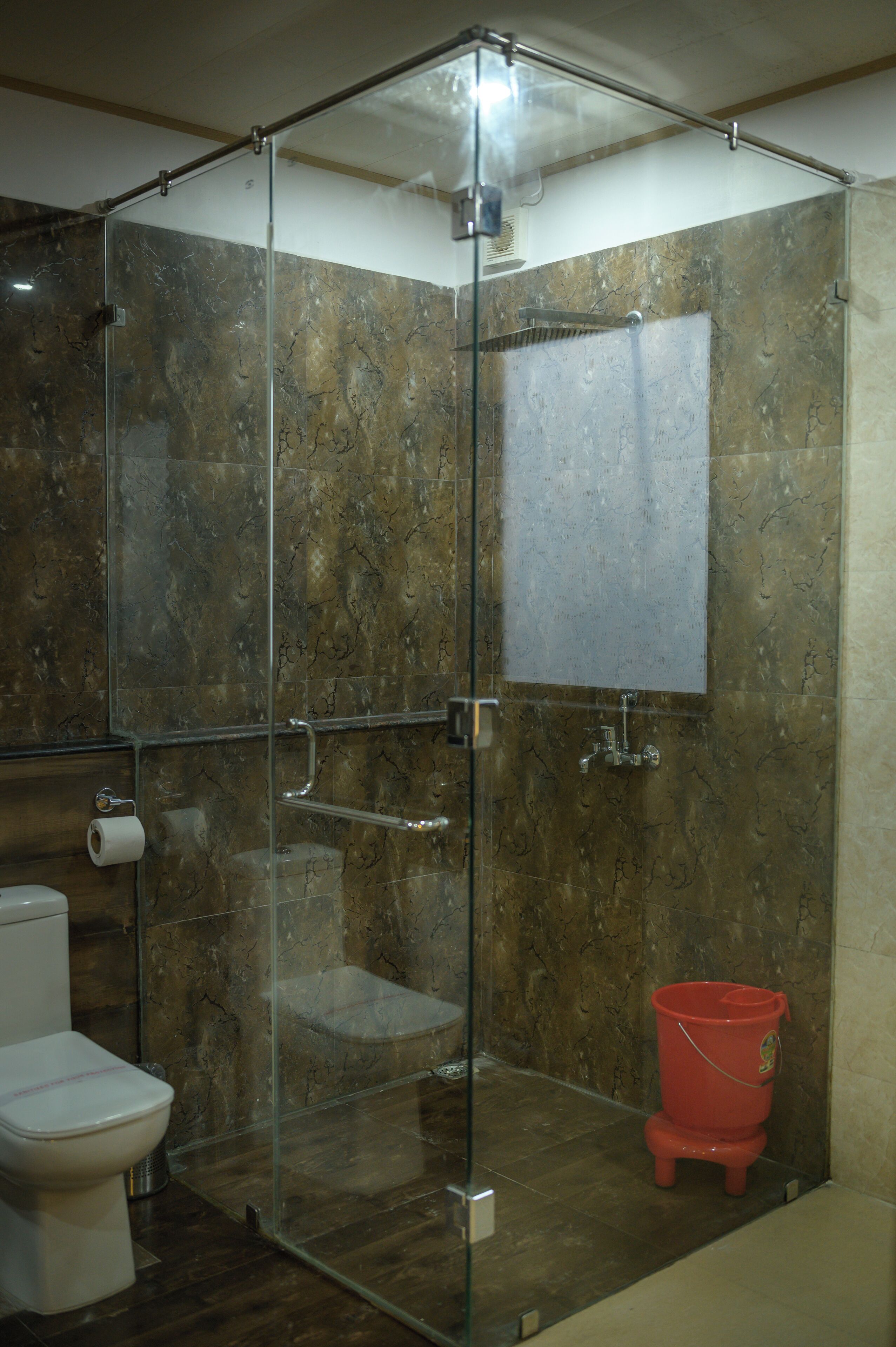Une cabine de douche fermée par des parois de verre, avec un mur en marbre, des toilettes et un seau rouge.