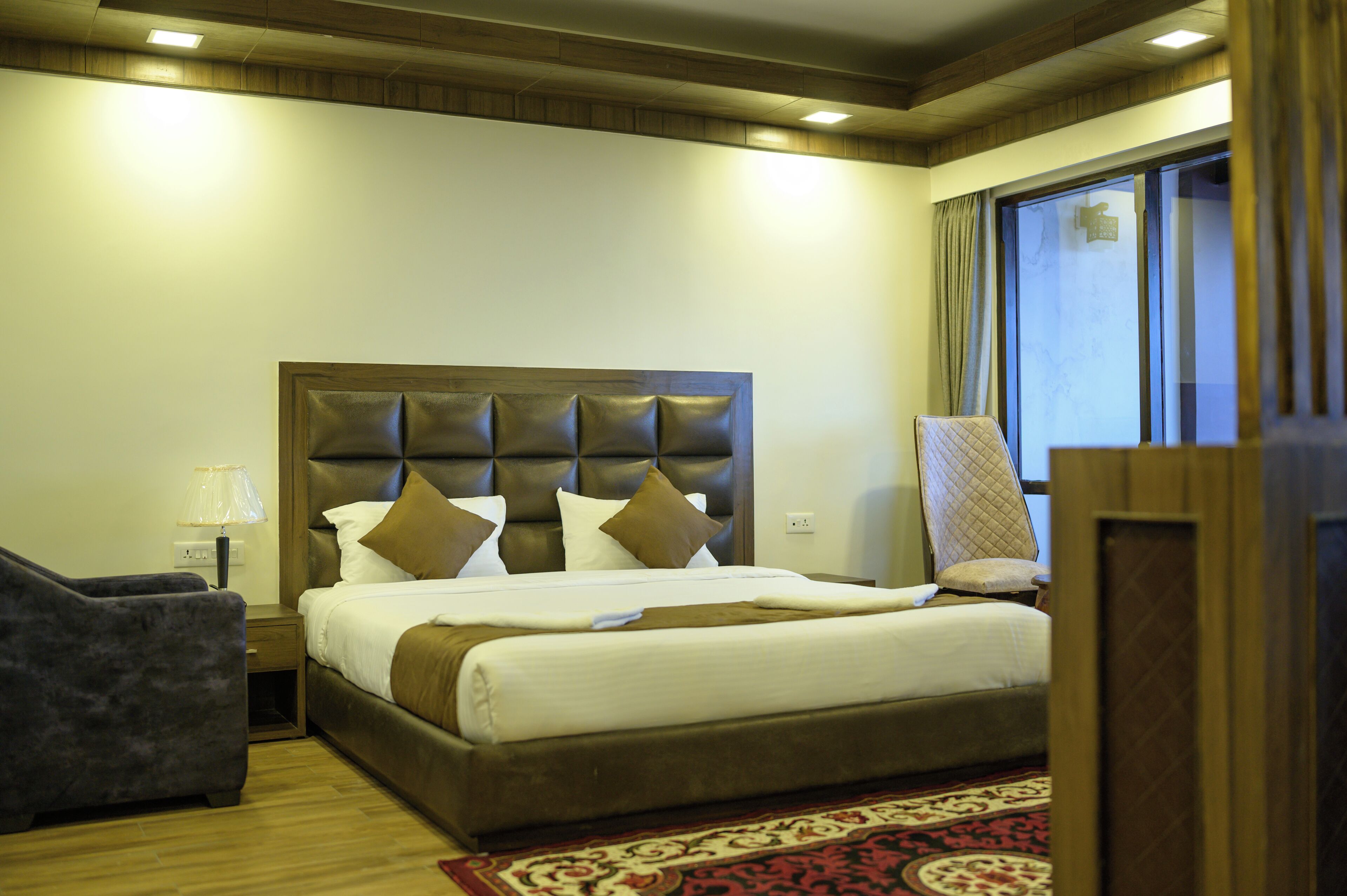 A hotel room with a large bed, a nightstand, a chair, and a window with curtains.