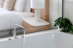 Et badeværelse med et hvidt badekar, en moderne armatur, et lille hvidt natbord med en lampe og en grøn plante.