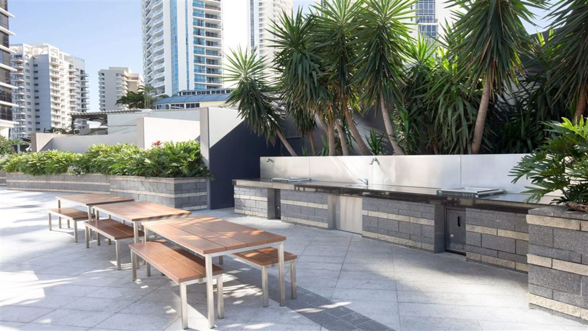 An outdoor patio with wooden tables and benches, a built-in barbecue grill, and lush greenery.