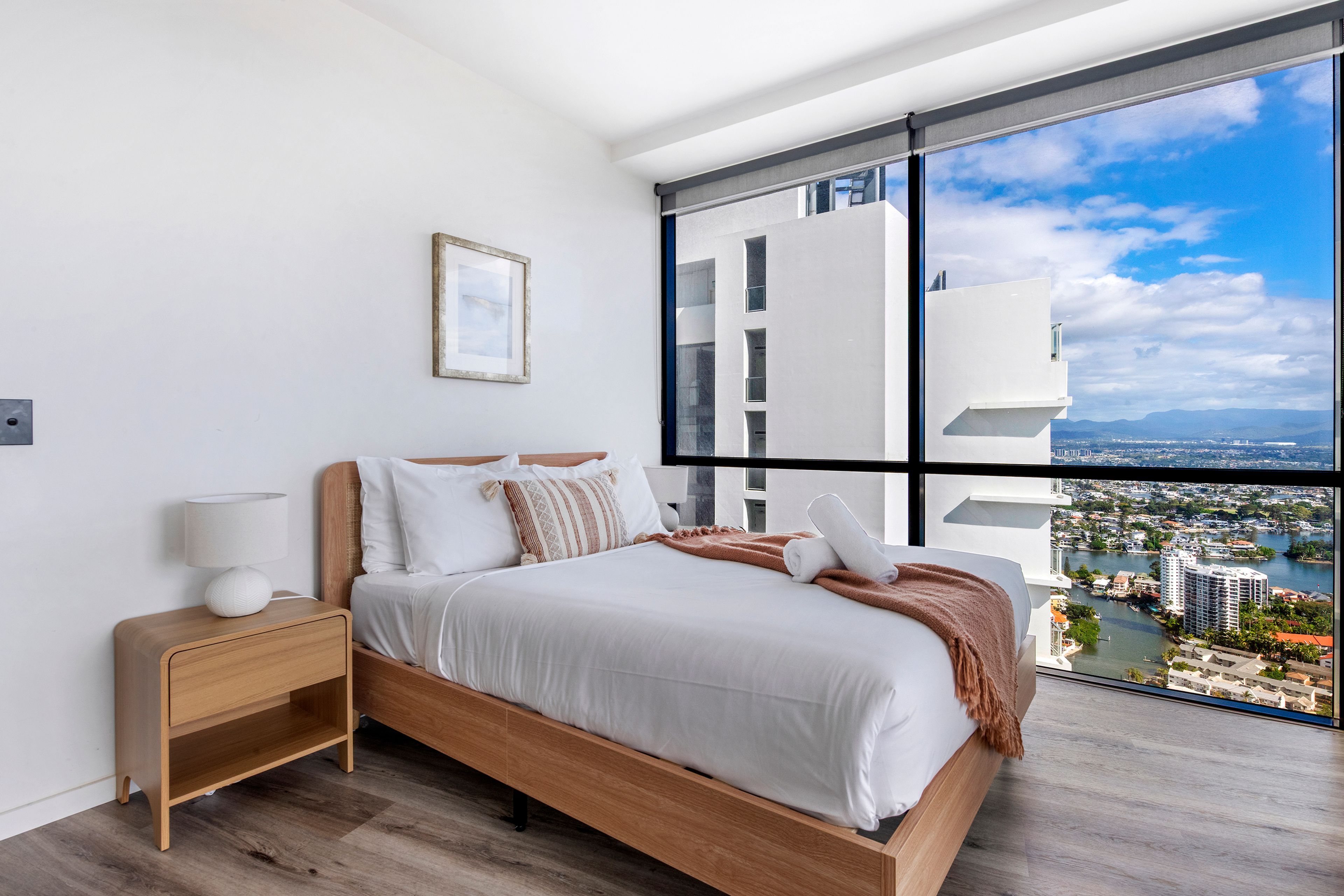 Un dormitorio moderno con un ventanal grande, una cama con ropa blanca, una mesita de noche y un cuadro enmarcado en la pared.