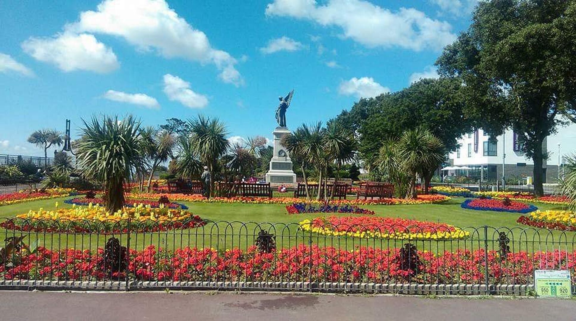 Un parc avec une statue, des parterres de fleurs et un bâtiment en arrière-plan.