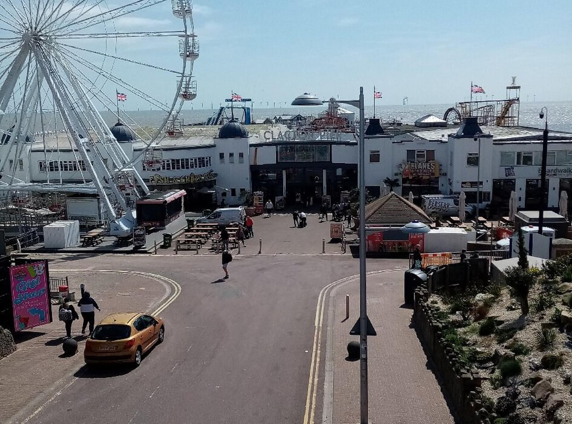 Uma vila costeira com uma roda-gigante imponente, um cais com barcos e diversos estabelecimentos comerciais e restaurantes.